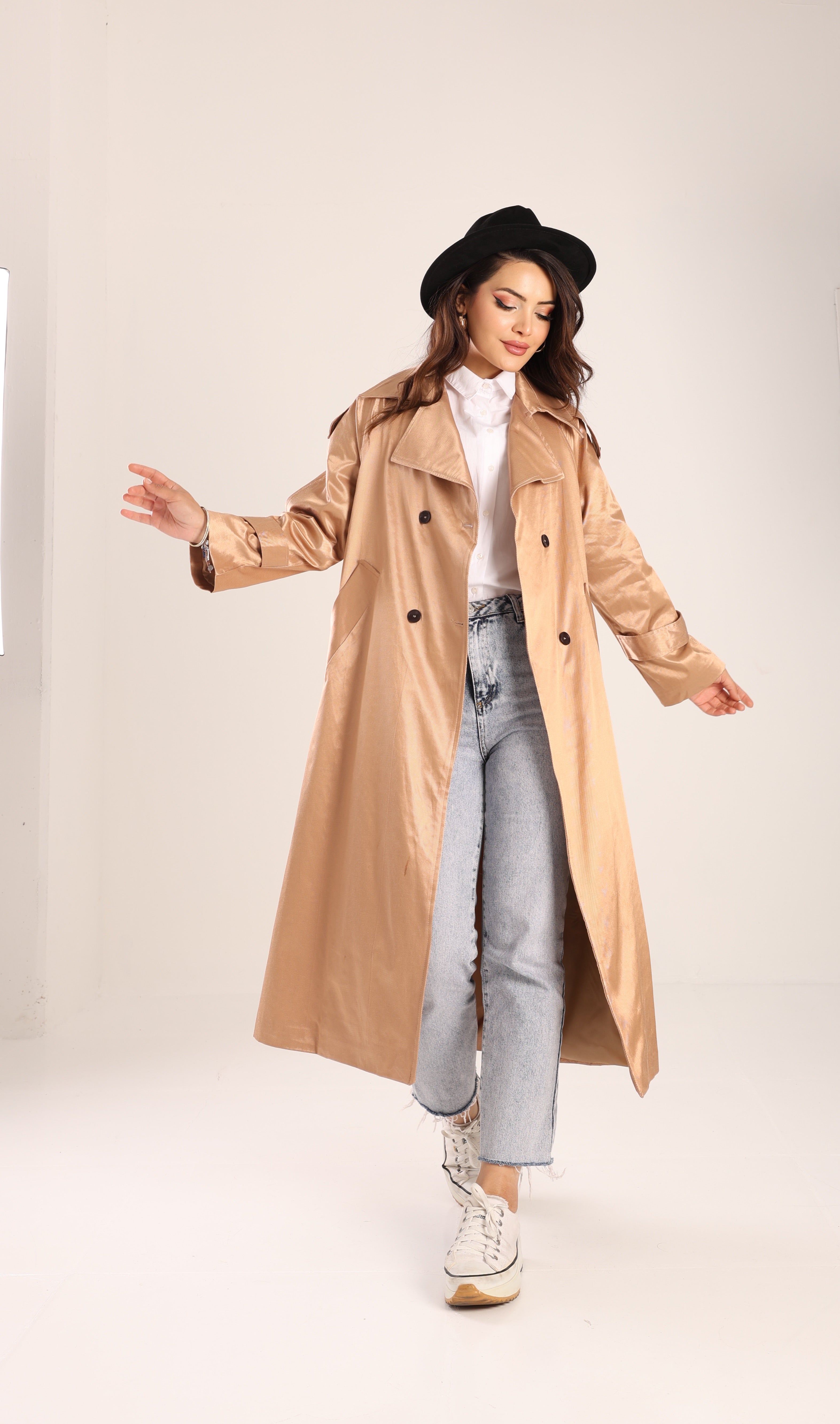 Woman wearing a beige trench coat, white shirt, gray jeans, and black hat on a white background